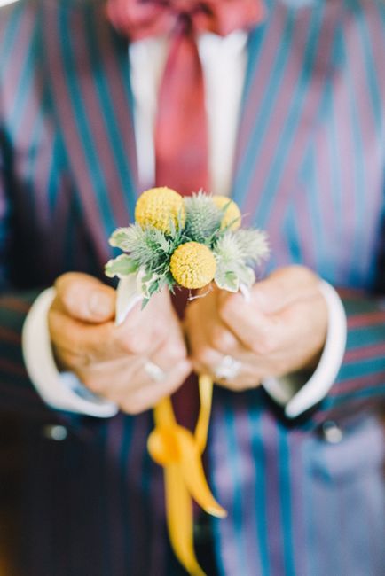 A person in a pinstripe suit and white shirt holds a small floral bouquet with both hands, positioned in front of their chest. The bouquet features yellow pom-pom-like flowers, light green foliage wit