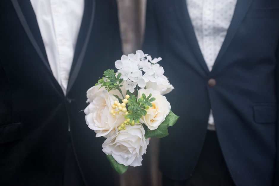 A close-up of a boutonnière floral arrangement featuring white roses, small white hydrangea blooms, and sprigs of greenery with yellow accents, held between the lapels of two men dressed in dark suit