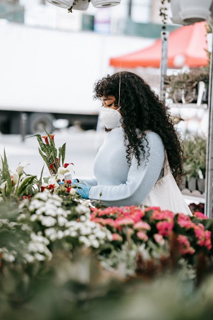 A woman with long, curly dark hair wearing a white face mask, light blue sweater, and gloves, selecting and arranging fresh floral bouquets at an outdoor flower market. The arrangement features a vari