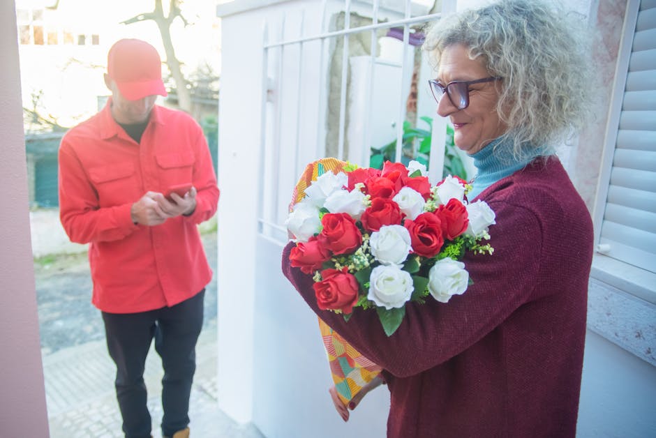 A woman with curly gray hair and glasses, dressed in a burgundy sweater and light blue turtleneck, is standing outside a house holding a vibrant bouquet of fresh roses, featuring red and white blooms