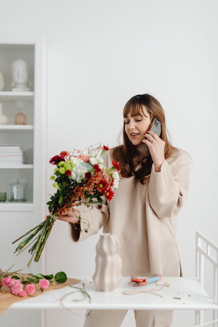 A young woman with shoulder-length brown hair and bangs, wearing a cream-colored sweater, is holding a vibrant, fresh bouquet of mixed flowers in her left hand while speaking on a smartphone held to h